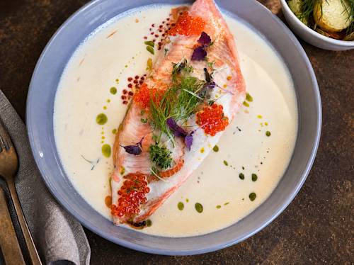 A plate with a poached salmon fillet in a creamy sauce, topped with fish roe and herbs, next to a bowl of roasted baby potatoes with dill.