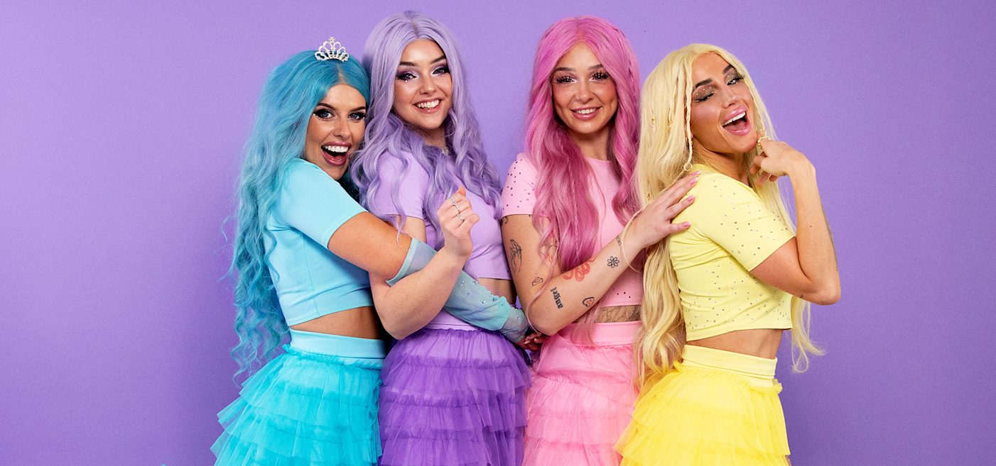 Four women with colorful wigs and matching outfits in shades of blue, purple, pink, and yellow stand smiling against a purple backdrop.
