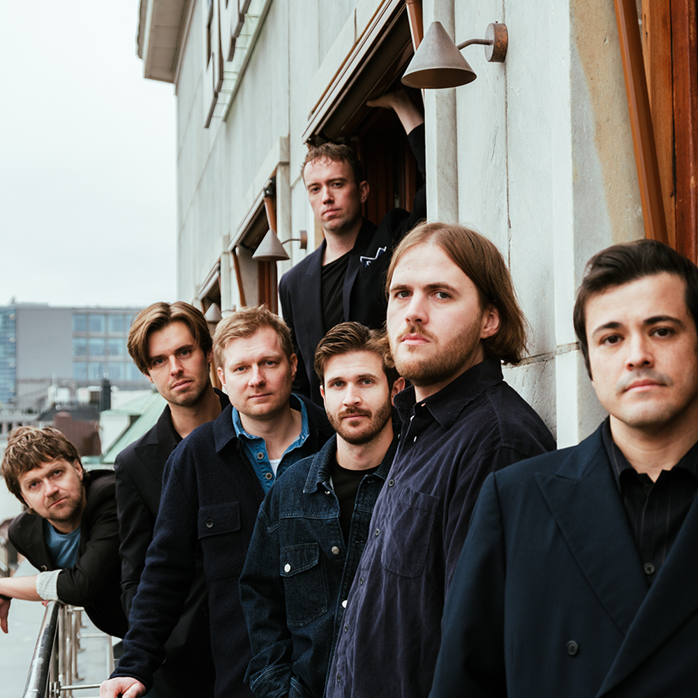 Sju män står i en rad utanför en byggnad och tittar mot kameran; i bakgrunden syns en del av ett stadslandskap.