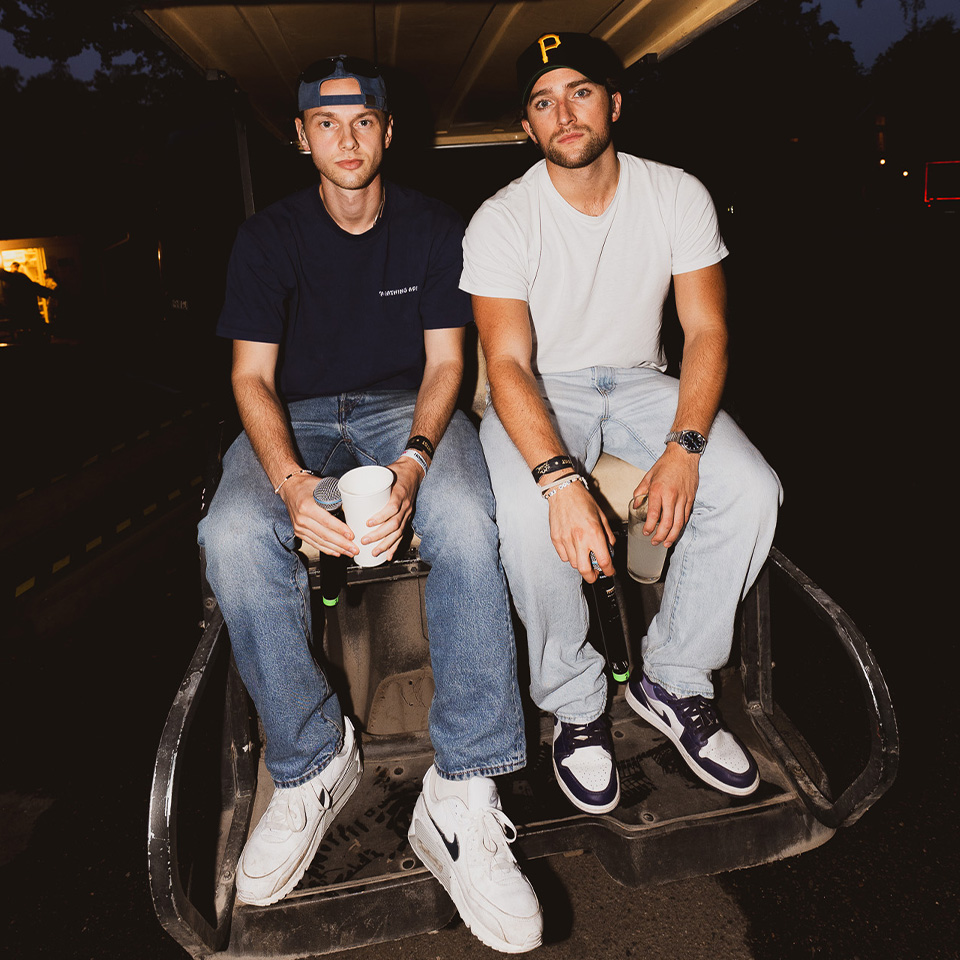 Två män sitter sida vid sida på baksidan av en golfbil på natten; båda bär fritidskläder, jeans, sneakers och basebollkeps, håller i drinkar och tittar in i kameran.