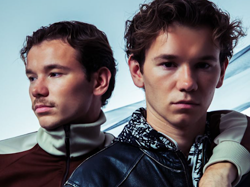 Two young men standing close together against a light background, one with a mustache and wearing a jacket, the other with curly hair and a patterned shirt.