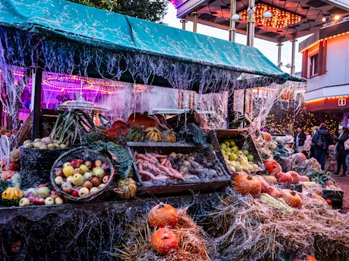 I ett marknadsstånd visas pumpor, äpplen och olika grönsaker täckta av falska spindelnät, med halmbalar och festliga lampor i bakgrunden.