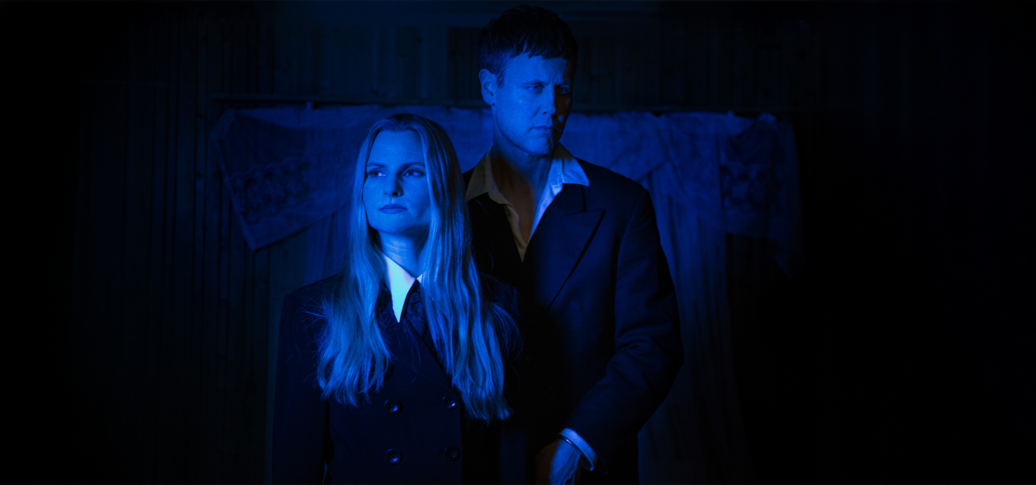 En kvinna och en man i mörka kostymer står sida vid sida, upplysta av blått ljus, med en svagt upplyst bakgrund.