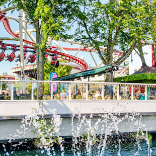 Människor sitter på ett utomhuscafé nära en fontän, med en rödvit berg- och dalbana och träd i bakgrunden.
