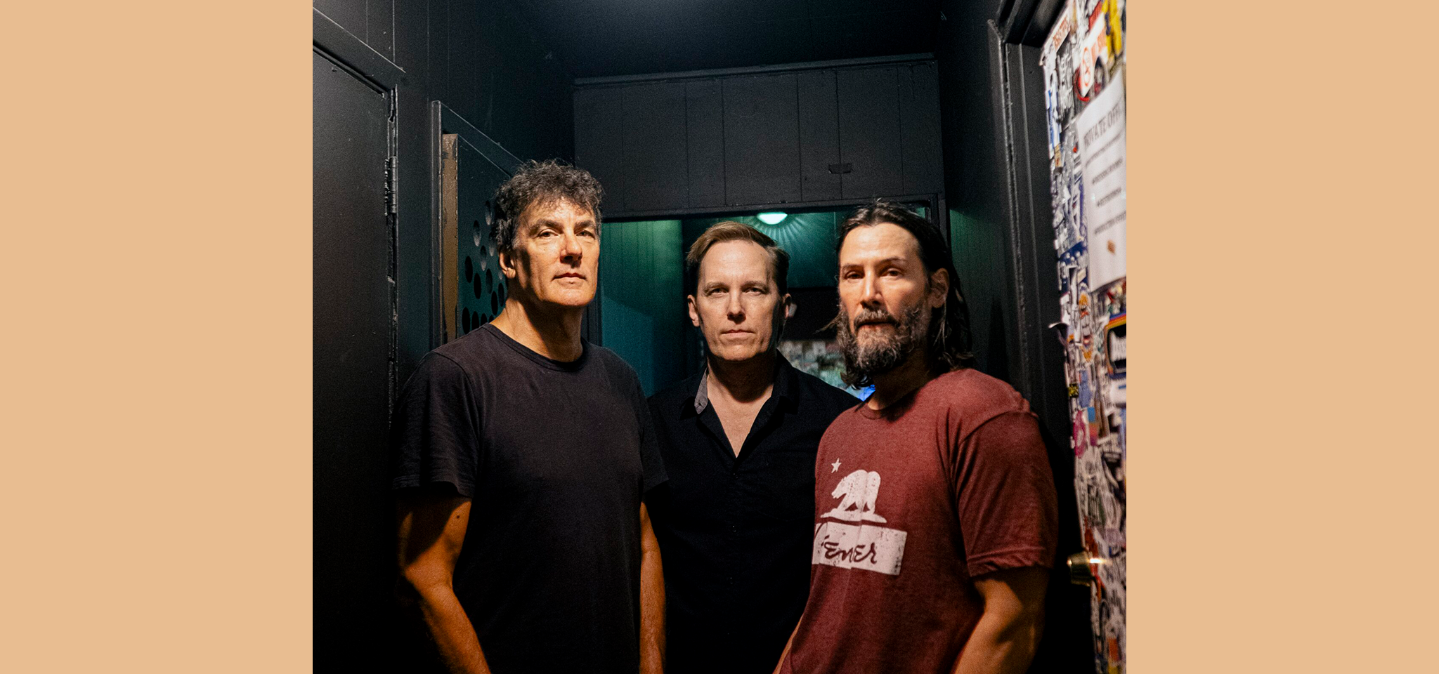 Tre män står tätt intill varandra i en svagt upplyst korridor och tittar rakt in i kameran; två bär mörka skjortor, en bär en röd T-shirt.