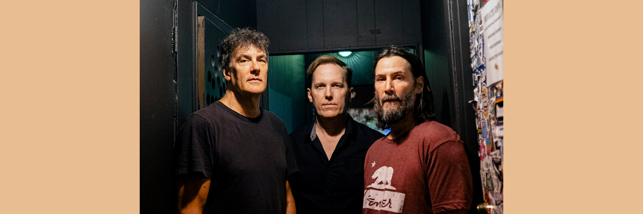 Tre män står tätt intill varandra i en svagt upplyst korridor och tittar rakt in i kameran; två bär mörka skjortor, en bär en röd T-shirt.