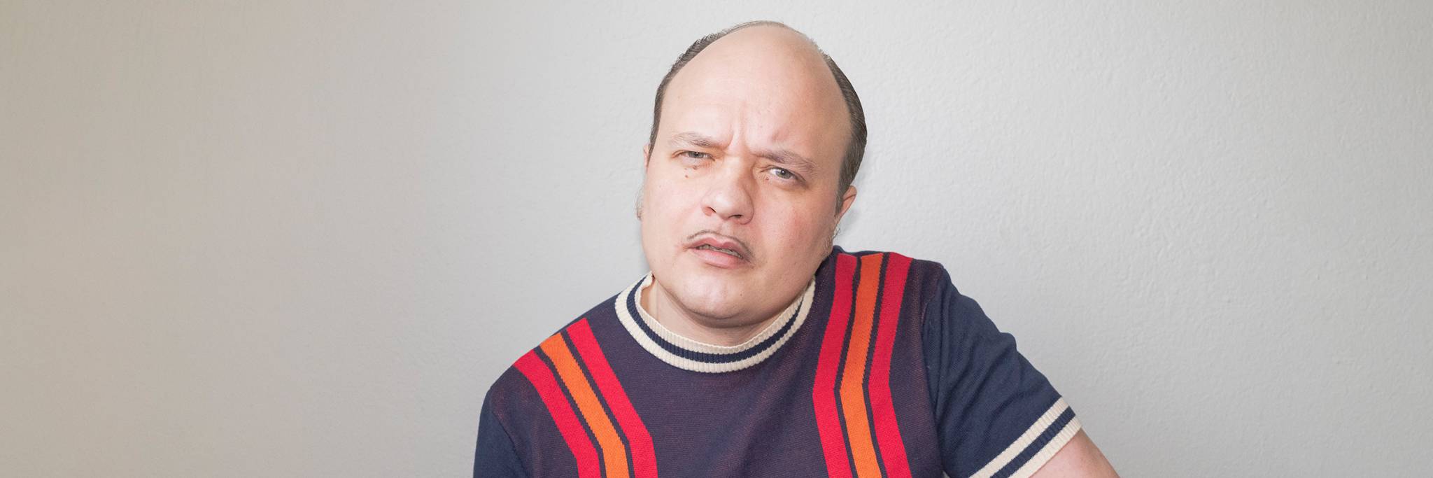 En man med tillbakadraget hårfäste och mustasch, klädd i en marinblå skjorta med röda ränder, sitter framför en vanlig ljus vägg och ser lite förbryllad ut.