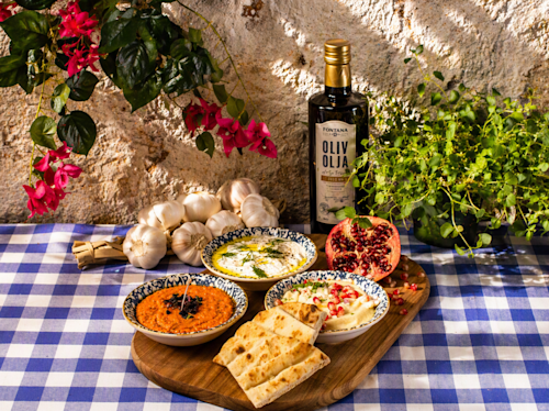 A wooden board with flatbread, three dips, garlic, pomegranate, a bottle of olive oil, and herbs on a blue checkered tablecloth, set outdoors with flowers and sunlight.
