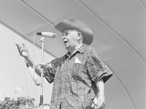 An elderly man in a patterned shirt and cowboy hat speaks into a microphone on an outdoor stage.