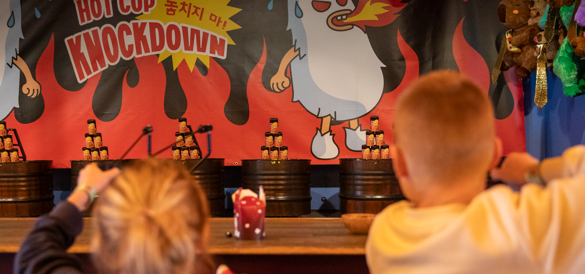 Två barn siktar på staplade burkar i ett tivoliliknande spelbås med en "Hot Cup Knockdown"-skylt och en tecknad kyckling i bakgrunden.
