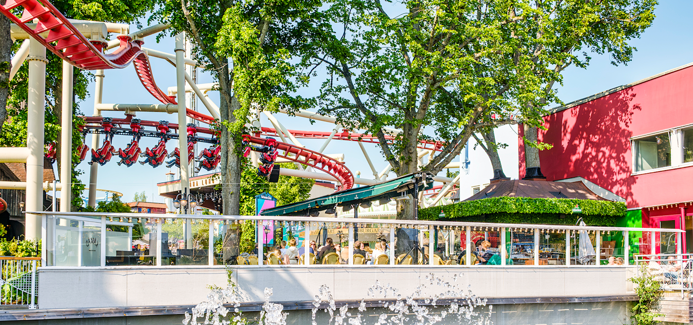A red roller coaster runs above trees and a patio seating area next to a red building, with people dining and water fountains in the foreground.