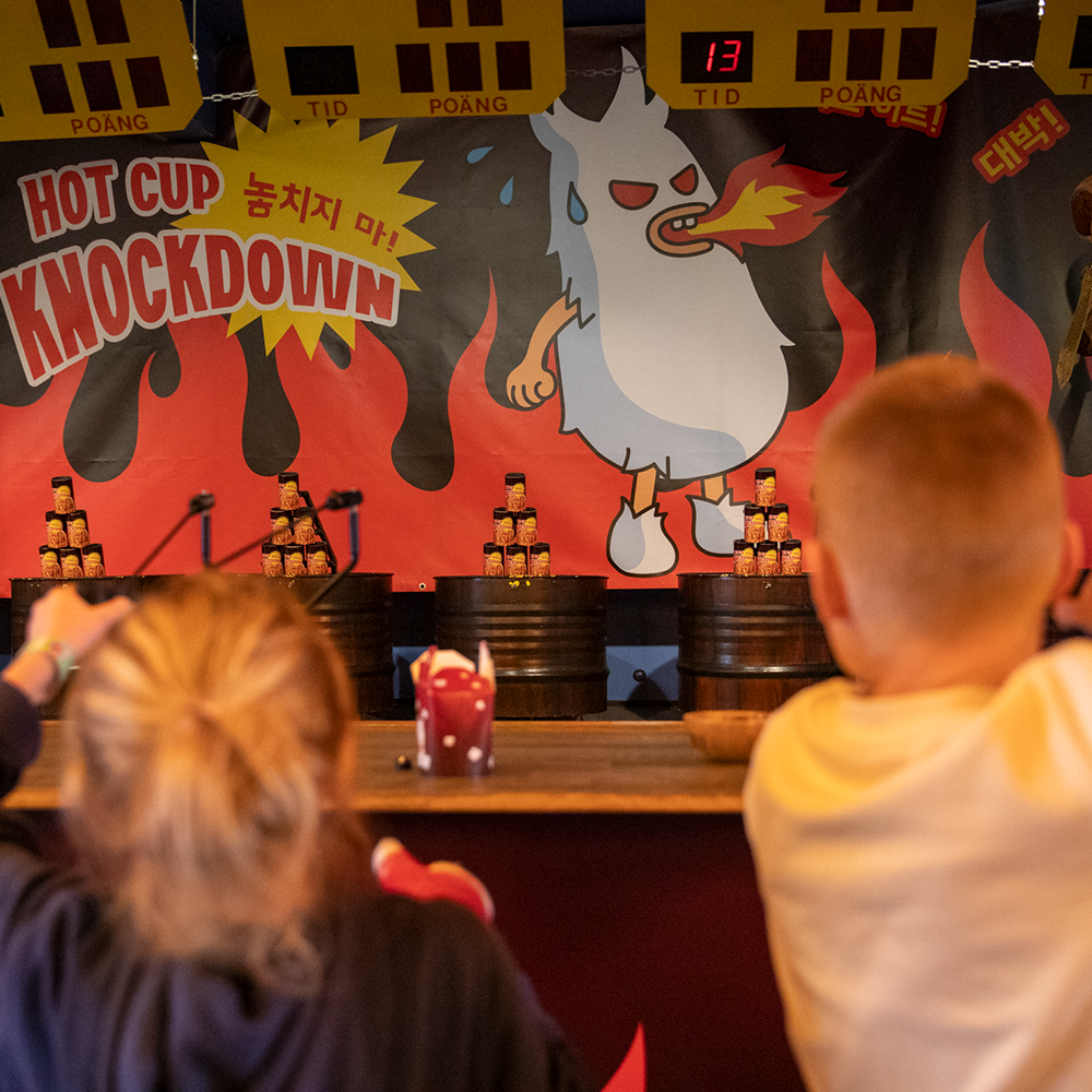 Två barn siktar med slangbellor på staplade muggar i ett tivolistånd som heter "Hot Cup Knockdown", med en tecknad figur som andas eld i bakgrunden.