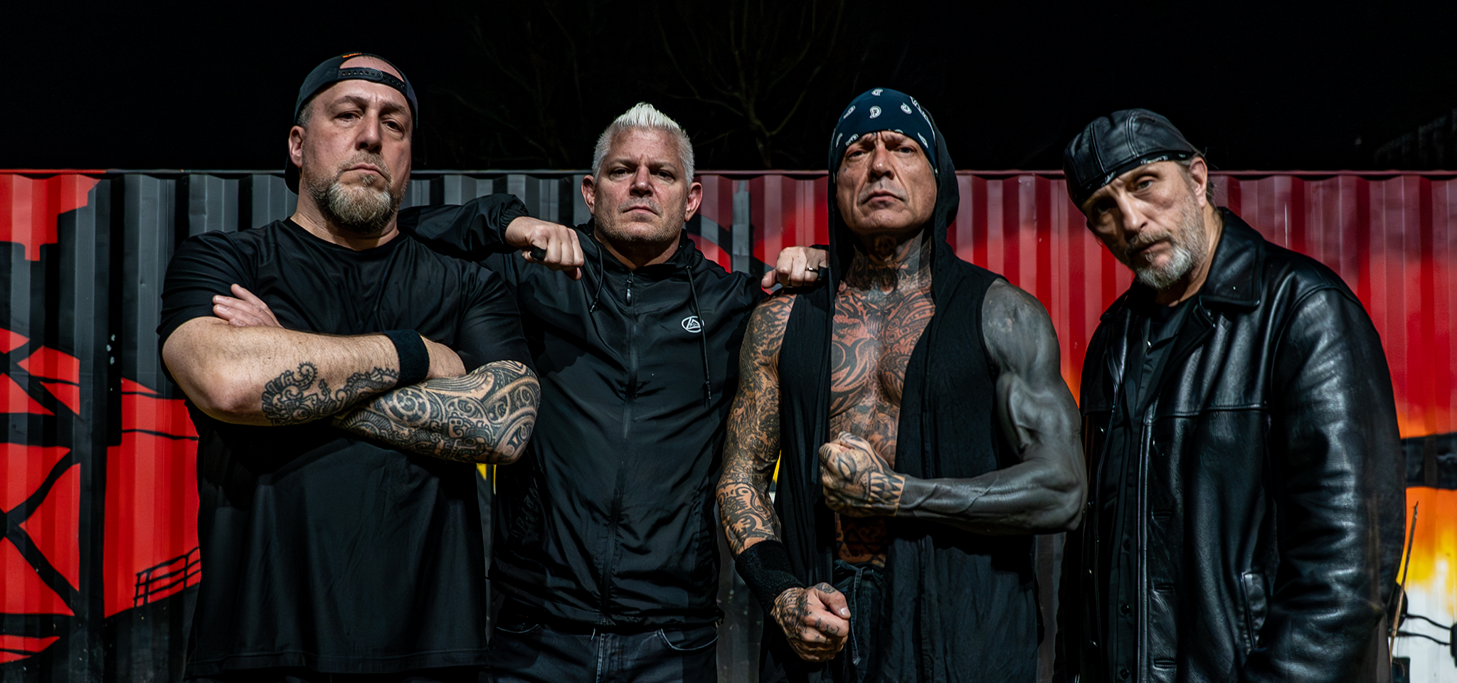 Fyra män med tatueringar och allvarliga ansiktsuttryck poserar framför en graffitiklädd vägg på kvällen.
