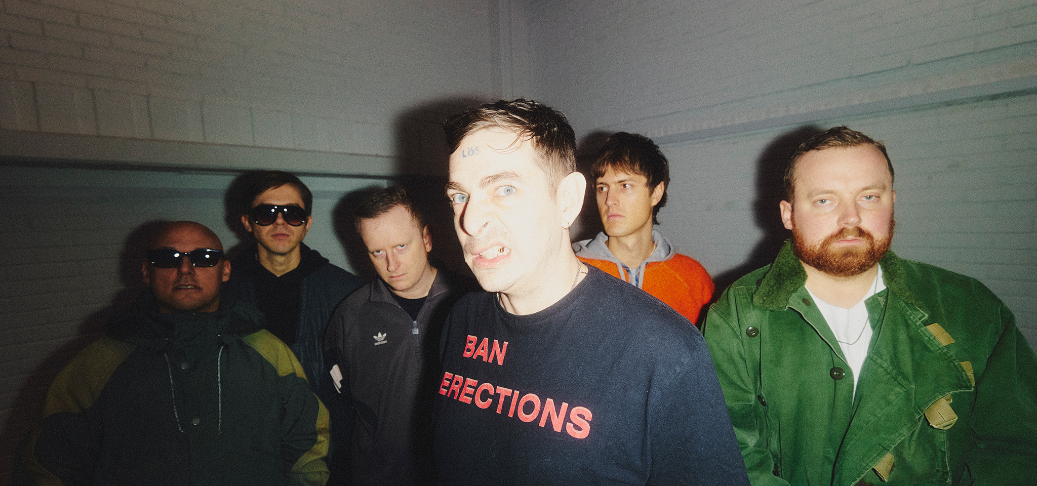Sex män står inomhus mot en vit tegelvägg, med en man längst fram iförd en svart tröja med texten "BAN ERECTIONS" i röda bokstäver och med ett förvridet ansiktsuttryck.