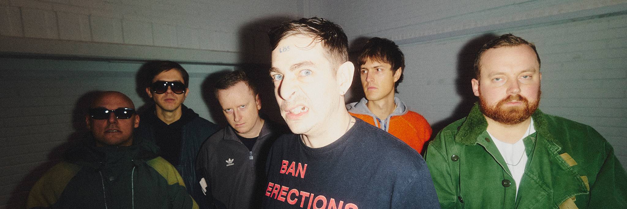 Six men stand together against a white brick wall. The man in front wears a black shirt with red text and makes a face, while the others look forward with neutral or serious expressions.