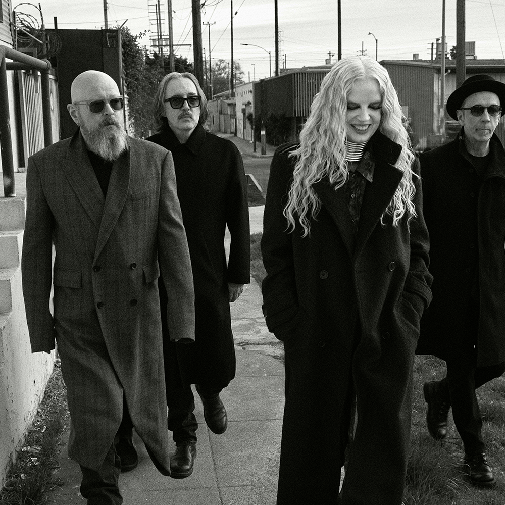 Fyra personer i långa rockar går tillsammans utomhus på en trottoar; bilden är i svartvitt.