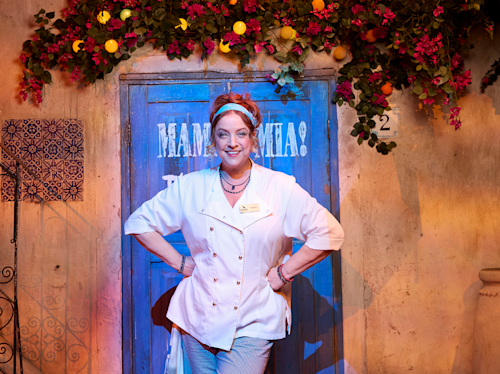 A woman in a chef's coat and headband stands smiling with hands on hips in front of a blue door decorated with flowers and the words "Mamma Mia!" painted on it.