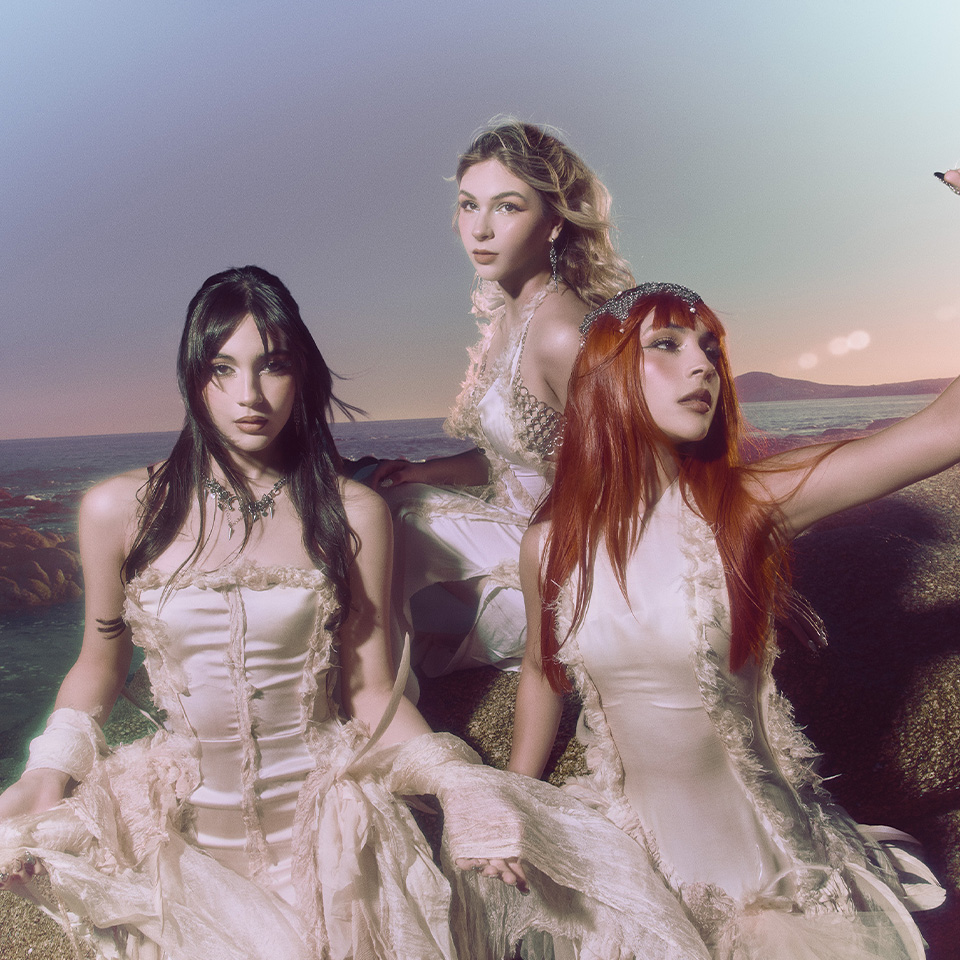 Three women in flowing, off-white dresses sit on rocks by the ocean at sunset, looking toward the camera.