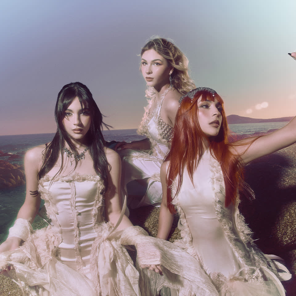 Three women in flowing, off-white dresses sit on rocks by the ocean at sunset, looking toward the camera.