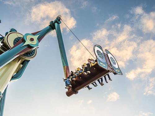 People are seated on a large swinging amusement park ride against a partly cloudy sky.