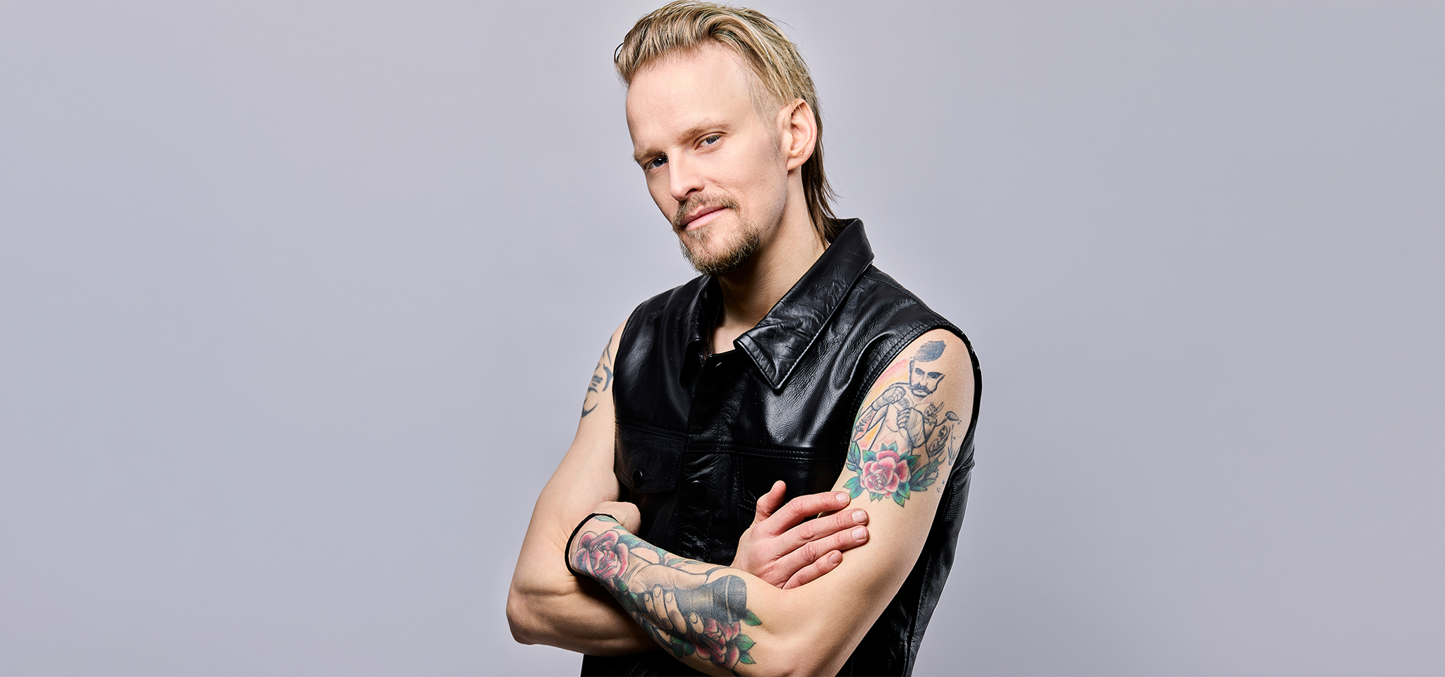 En man med ljust hår och vältrimmat skägg bär en ärmlös svart läderväst, visar tatuerade armar och står med armarna i kors mot en ljus bakgrund.