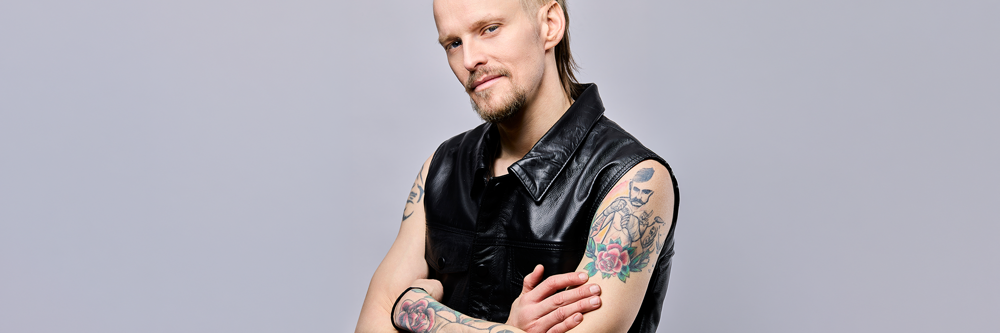 En man med ljust hår och vältrimmat skägg bär en ärmlös svart läderväst, visar tatuerade armar och står med armarna i kors mot en ljus bakgrund.