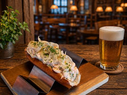 A lobster roll topped with chopped herbs and onions on a wooden board, next to a glass of beer in a dimly lit restaurant setting.