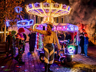 En utklädd artist sjunger i en mikrofon bredvid clowner och en folkmassa på en starkt upplyst karneval med en karusell i bakgrunden på natten.