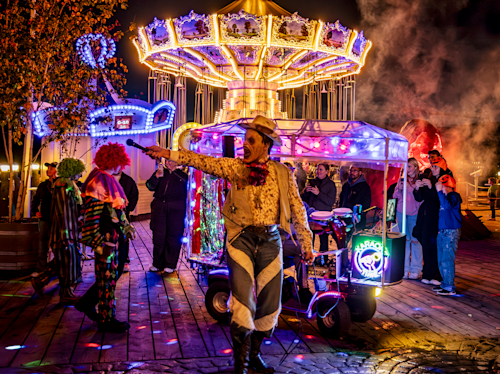 En utklädd artist sjunger i en mikrofon bredvid clowner och en folkmassa på en starkt upplyst karneval med en karusell i bakgrunden på natten.