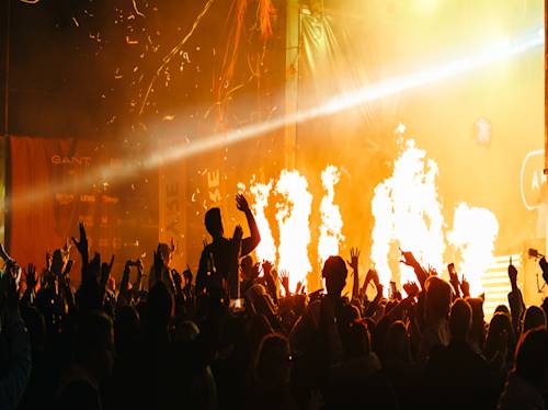 En stor publik på en konsert med uppsträckta händer, i siluett mot ljusa flammor och scenljus.