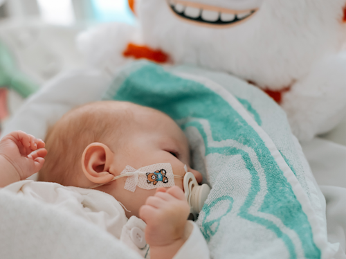 En baby med napp och ett tecknad bandage på kinden ligger på en säng, delvis täckt av en blågrön och vit filt, nära en plyschleksak med ett stort leende.