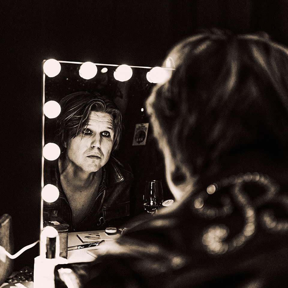 A person with tousled hair looks at their reflection in a brightly lit makeup mirror, wearing a leather jacket, with makeup items and a drink visible on the table.