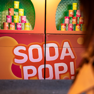 Färgglada läskburkar staplas på hyllor ovanför en skylt med texten "SODA POP!" i feta vita och rosa bokstäver.