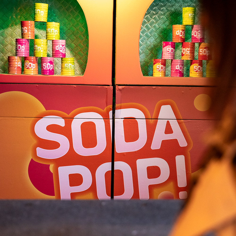 Färgglada läskburkar staplas på hyllor ovanför en skylt med texten "SODA POP!" i feta vita och rosa bokstäver.