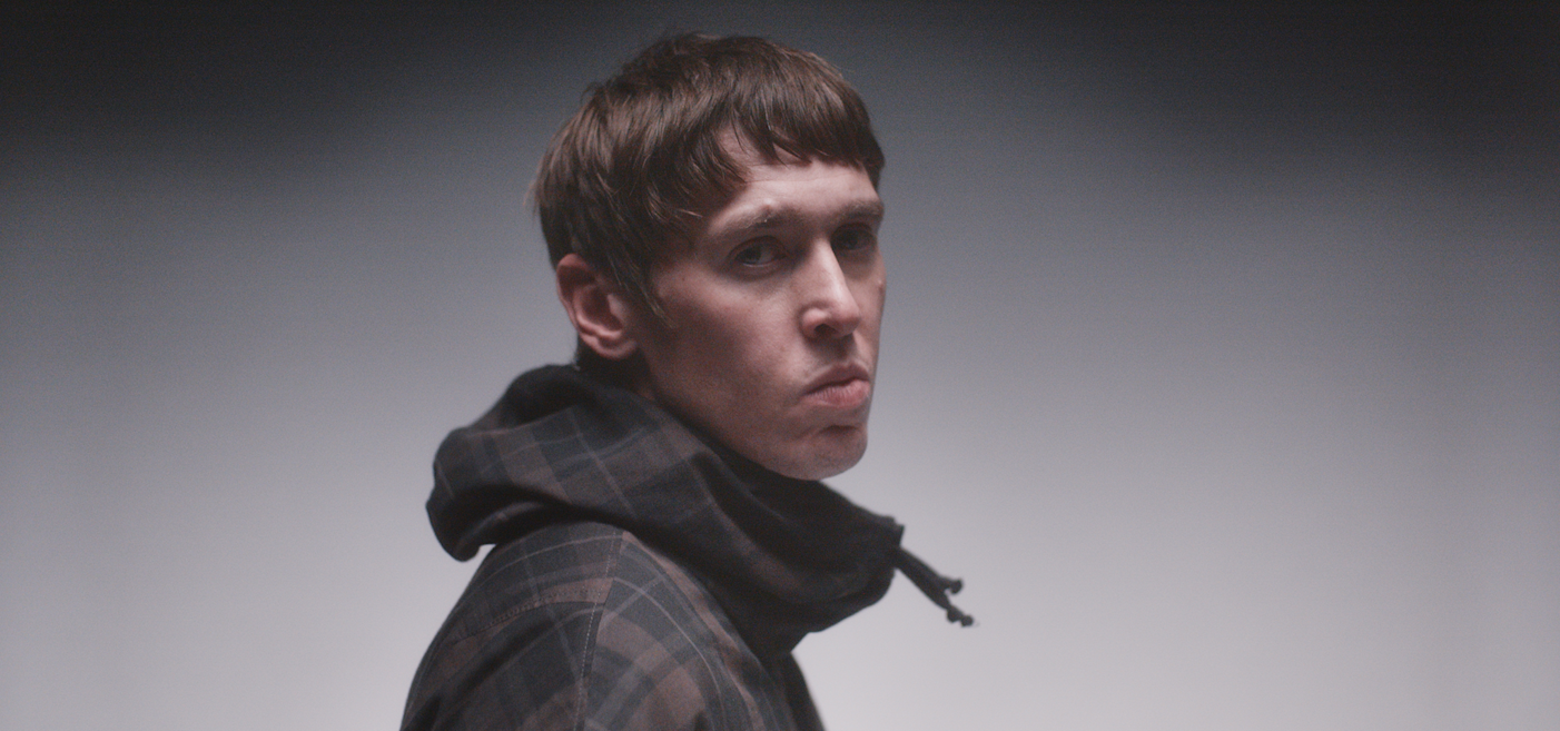 En ung man med kort brunt hår och rutig jacka står mot en enfärgad bakgrund och tittar lite åt sidan.