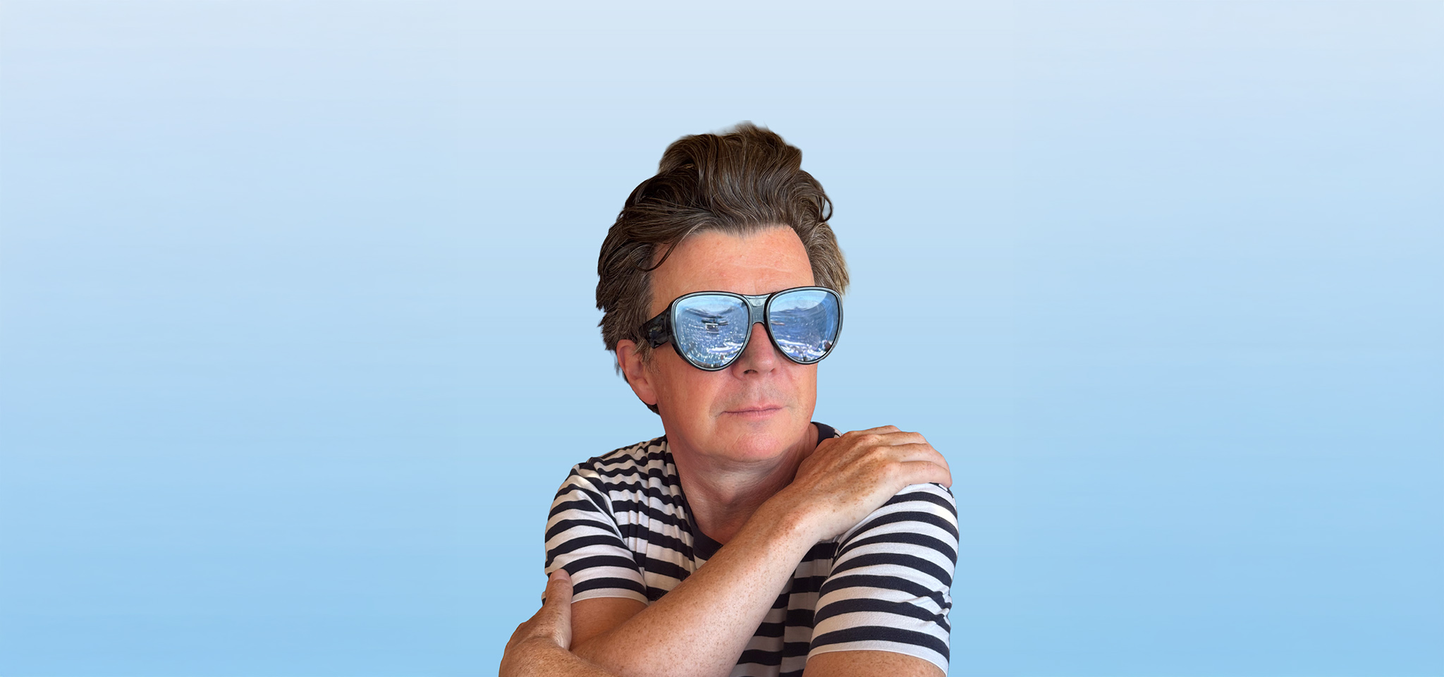 Person i randig skjorta med stora solglasögon, med blå himmel och vatten som reflekteras i glasen, poserar mot en blåtonad bakgrund.