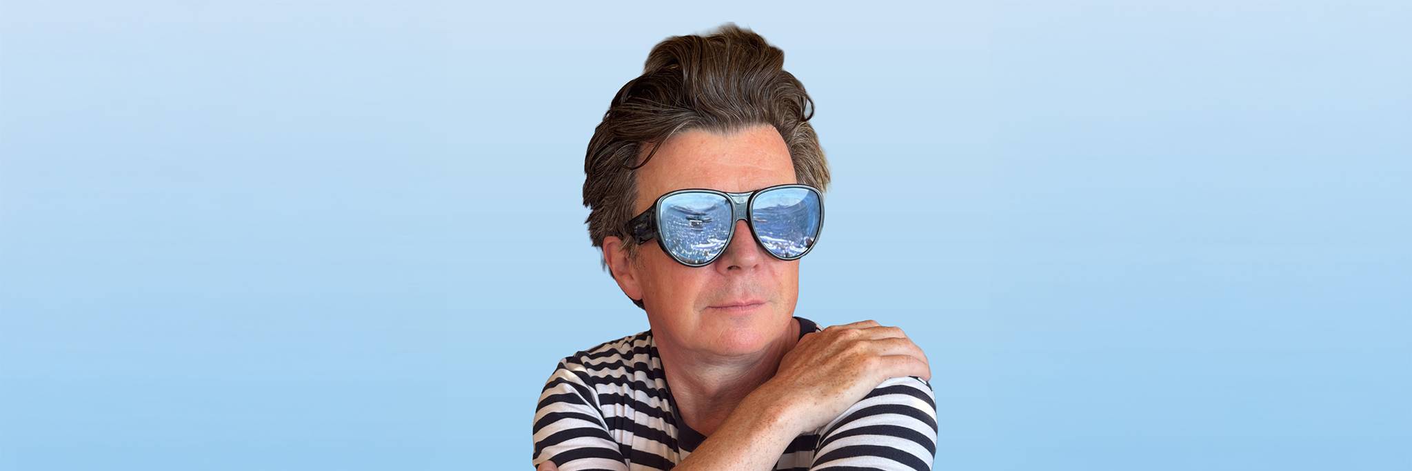 Person i randig skjorta med stora solglasögon, med blå himmel och vatten som reflekteras i glasen, poserar mot en blåtonad bakgrund.