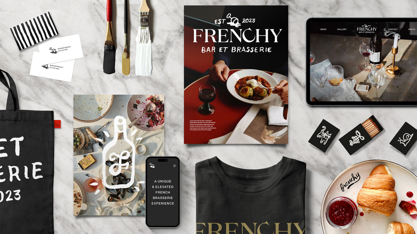 An overview of Frenchy branded assets laying on a marble table, included a magazine, match boxes, dinner ware, paintbrushes, a tote bag, and more.