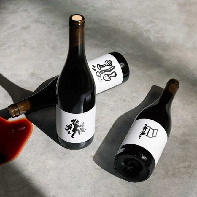 3 Frenchy branded wine bottles with different illustrations on their labels, one of the bottles is knocked over with red wine spilling out.