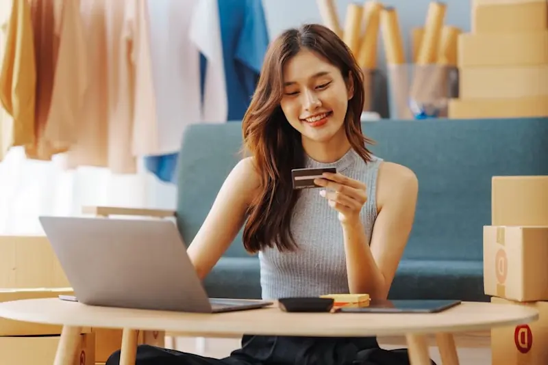 Smiling woman holding a credit card while shopping online with a laptop, surrounded by boxes in a cozy home setting.