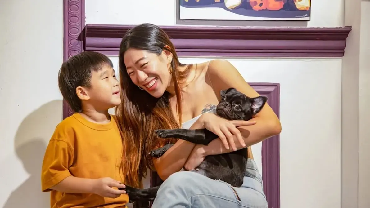 Smiling woman holding a black pug while sitting next to a young boy at What The Pug café in Singapore, enjoying a playful moment.