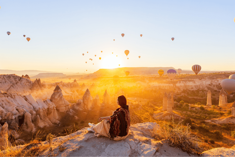 A woman sitting on a rocky cliff, watching hot air balloons rise over the fairy chimney rock formations at sunrise in the magical Cappadocia.