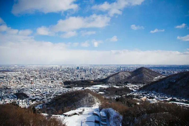Panoramic winter view of Sapporo from Okurayama Ski Jump Stadium, with snowy rooftops, rolling hills, and a bright blue sky.