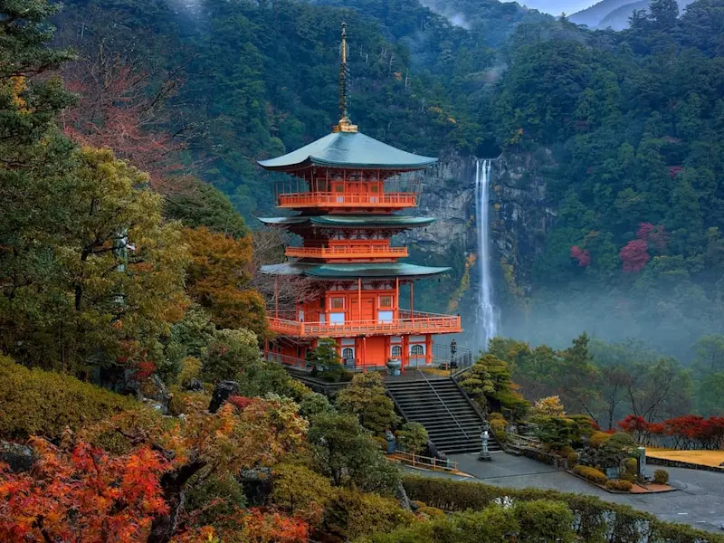 Three-tiered Seiganto-ji pagoda with Nachi Falls cascading behind it, surrounded by lush forest in autumn near Wakayama, Japan.