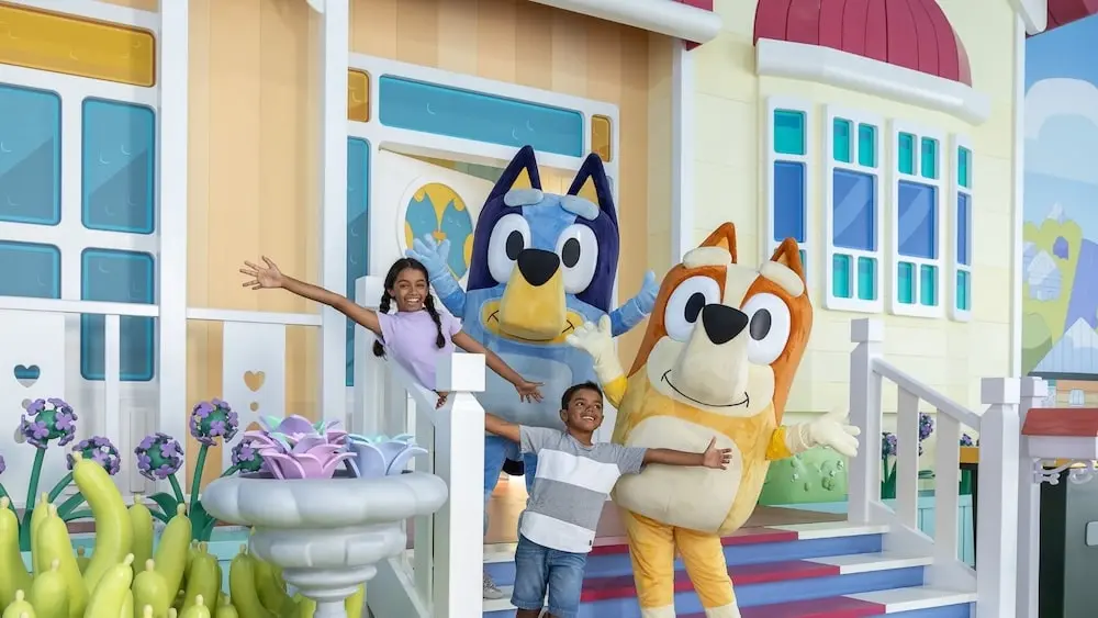 Two dark-skinned children reaching out and posing with Bluey and Bingo character performers in front of a replica of the Heeler Home at Bluey's World in Brisbane, Queensland.