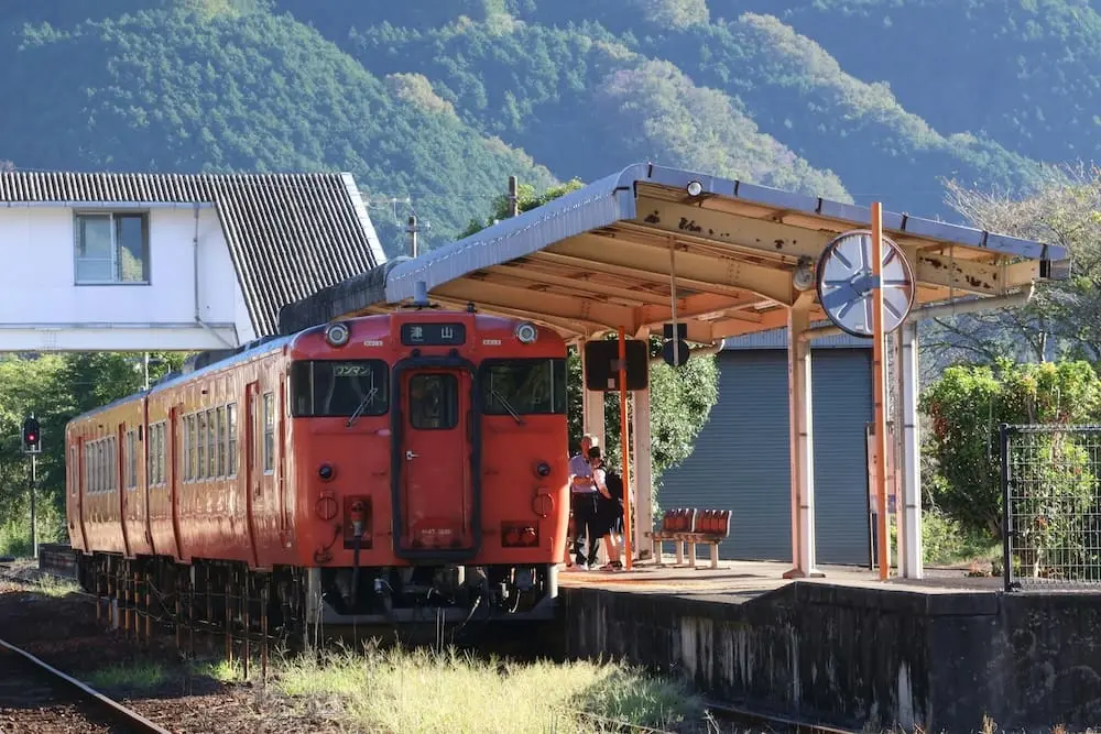 Red local train stopped at a rural platform in Tsuyama Station, Okayama, Japan, with forested hills and clear skies in the background.
