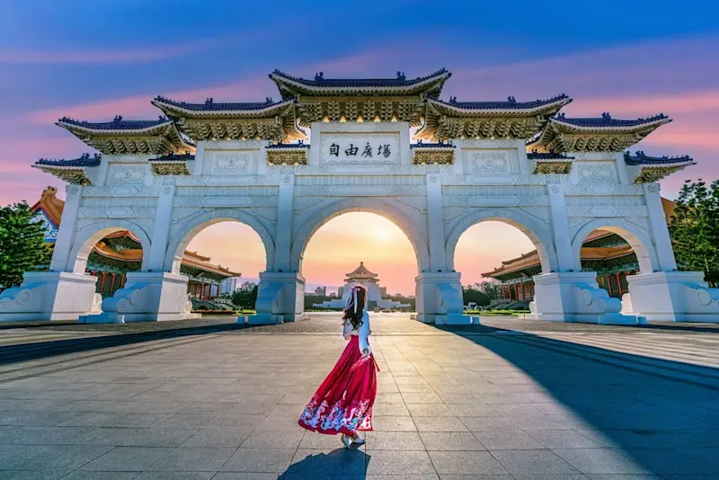 An Asian woman wearing a traditional Chinese dress walking towards the Archway at Chiang Kai-shek Memorial Hall in Taipei, Taiwan, with the sun setting visible through the arch.