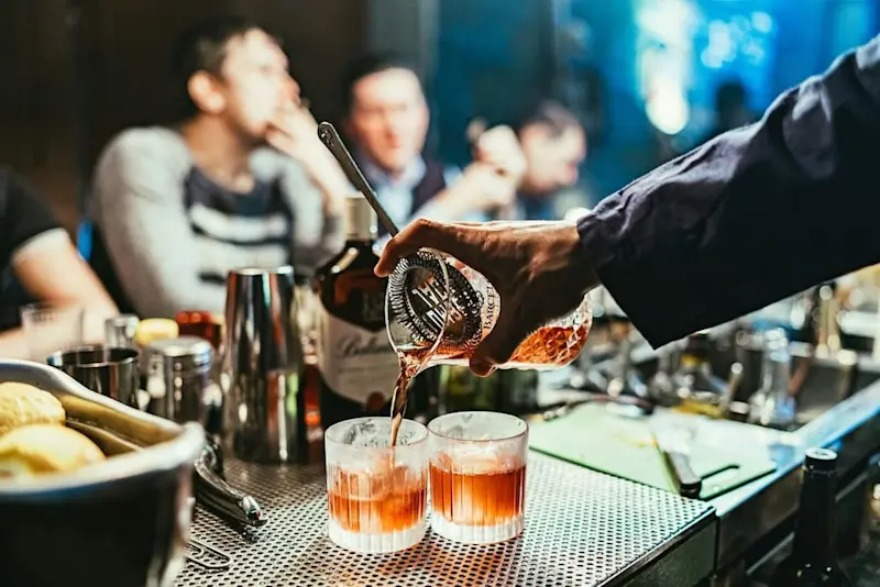 A bartender pouring whisky-colored alcohol into two glasses with ice at a busy bar.
