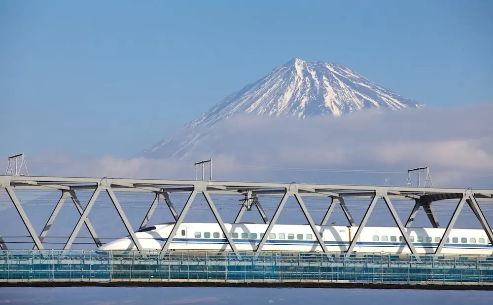 A blue-accented Shinkansen bullet train crossing a bridge in Shizuoka with snow-streaked, cloud-covered Mount Fuji in the background.