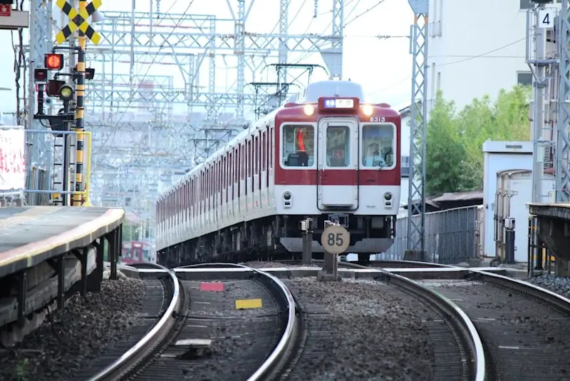 A red and white Kintetsu train approaches a station on curved tracks, framed by signals, platforms, and overhead power lines.